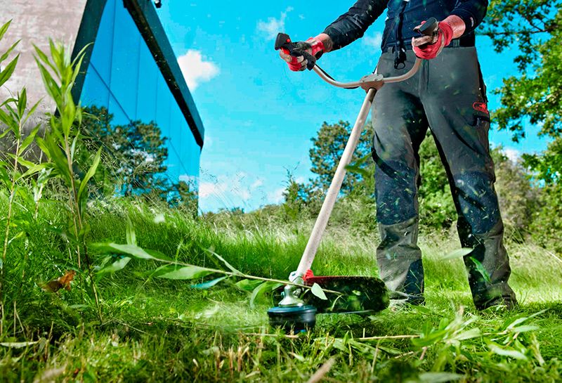 Человек косит высокую траву бензокосой WORCRAFT WGT52-280 возле дома.