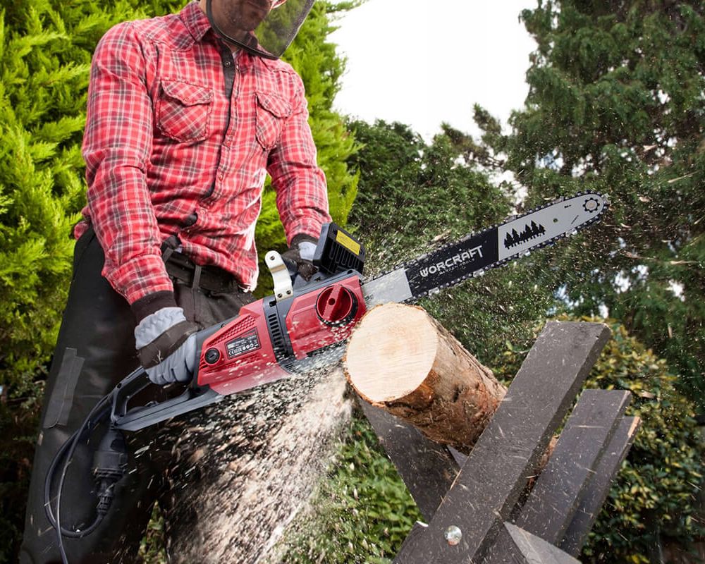 Людина в червоній сорочці розпилює колоду за допомогою електропили WORCRAFT WCE-2616 у саду.